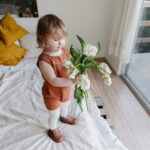 Cute toddler in rustic outfit holding tulips by a sunny window indoors.